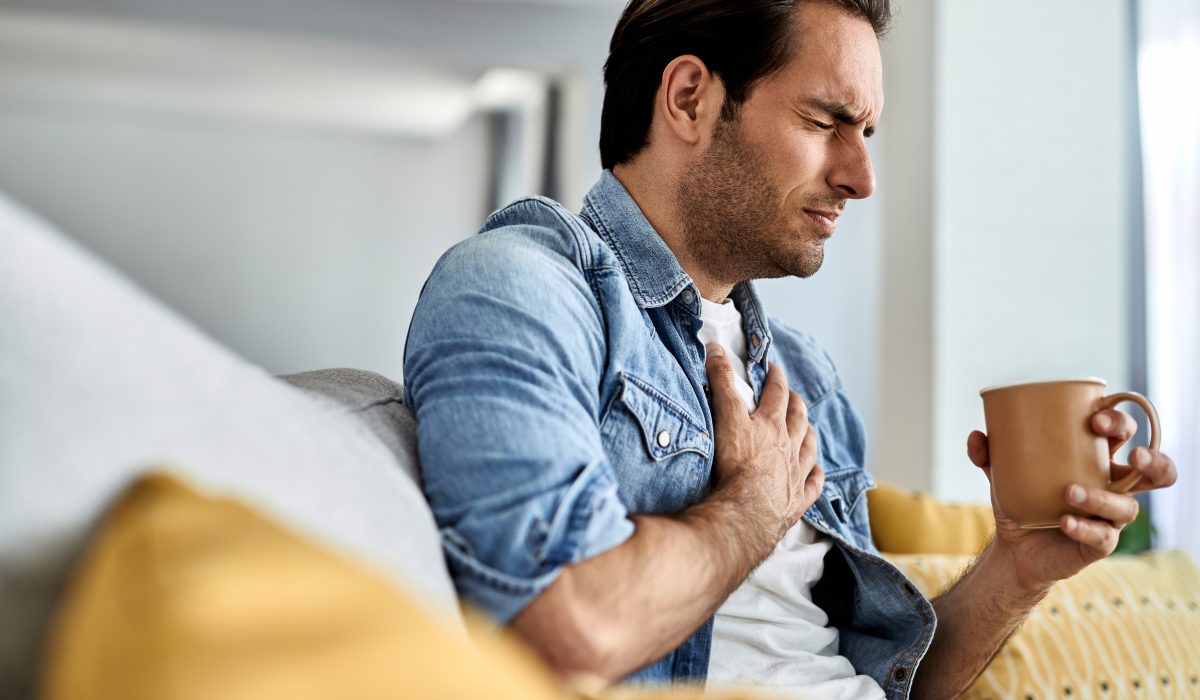 Young man feeling sick and holding his chest in pain while drinking tea in the living room.